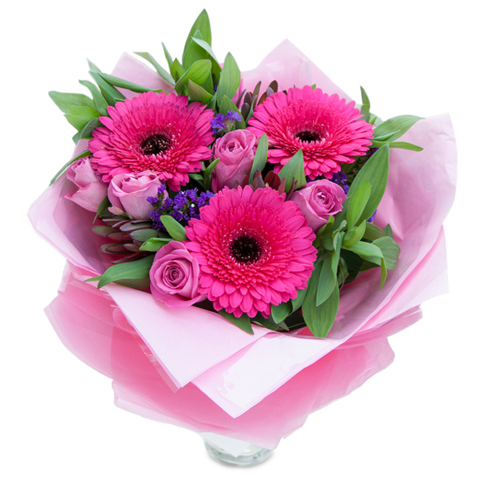 Pink and lavender tones mingle in this hand-styled bouquet to create a feeling of garden-fresh charm, anchored by three brilliant fuchsia Gerbera daisies whose radiating petals and dark centres give the arrangement a lively focal point. Complementing them are several roses in muted lavender-pink and dusty-rose shades, their petals rolled tightly at the centre and softening outward in velvety layers that speak of tenderness and romance. Between the larger blooms, clusters of deep purple statice provide texture and a cool visual counterbalance, like wildflower sparks tucked into a cultivated posy. Generous green foliage-suggestive of salal or eucalyptus-builds volume and a fresh, leafy scent that grounds the palette. The bouquet is wrapped as a gift: a soft, matte blush tissue underlies a sheer, slightly crinkled fuchsia-pink cellophane that catches the light and reinforces the pink theme; the folds are deliberate and neat, showing careful florist technique. At the base, the clear glass vase rim is visible, signalling this composition is arranged for immediate display on a mantel, bedside table or dining surface. In the quiet light of an early afternoon in Wood Green, near Alexandra Park and Green Lanes, the colours feel particularly vivid and uplifting. The olfactory impression is understated-roses lending a light perfume while gerberas give a faint, green sweetness-making this bouquet an ideal choice for birthdays, Mother's Day, anniversaries or a thoughtful delivery to brighten a neighbour's day. Crafted with local care, it reads as both joyful and intimate, perfectly suited to the neighbourhood's moments of celebration or comfort.